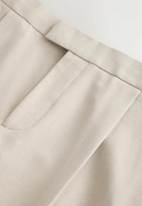 Gros plan sur un pantalon en tissu beige montrant la ceinture, les passants et les détails de la couture frontale sur un matériau lisse.