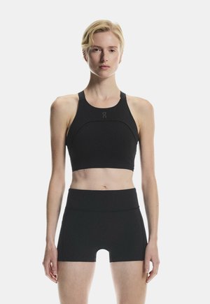 Blonde Person trägt schwarzen ärmellosen Crop Top und hoch taillierte Shorts, steht vor einem einfarbig weißen Hintergrund.