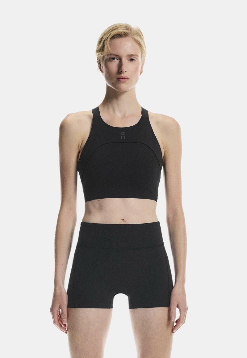 Personne blonde portant un crop top noir sans manches et un short taille haute, debout devant un fond blanc uni.