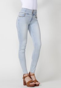 Jeans de tiro alto en azul claro con bolsillos decorativos, cierre de botón y diseño ajustado. Combinados con sandalias de plataforma marrones.