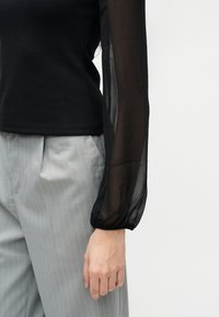 Άτομο που φοράει μαύρο τοπ με μακριά μανίκια και διάφανα μανίκια, καθώς και ανοιχτό γκρι παντελόνι με λεπτές ρίγες, στέκεται μπροστά σε λευκό φόντο.