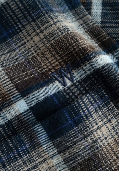 Tissu en tartan en laine présentant des motifs de carreaux bleu foncé, marron et crème, avec un accent brodé "W" en fil bleu. Texture douce.