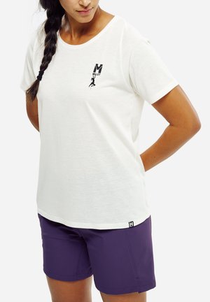 Femme aux cheveux tressés portant un T-shirt blanc Millet avec un logo d'escalade et un short violet, debout les mains derrière le dos.