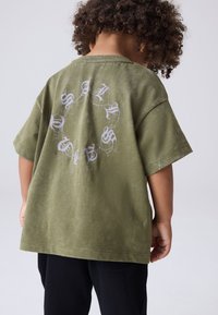 Barn med lockigt hår, sett bakifrån, bär en olivgrön T-shirt med vit gotisk text som bildar en cirkel på ryggen, kombinerad med svarta byxor.