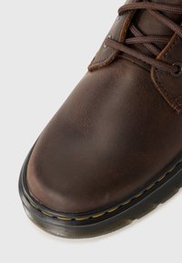Botte en cuir marron avec une finition lisse, bout rond, lacets marron et une semelle en caoutchouc noir accentuée de coutures jaunes le long du bord.