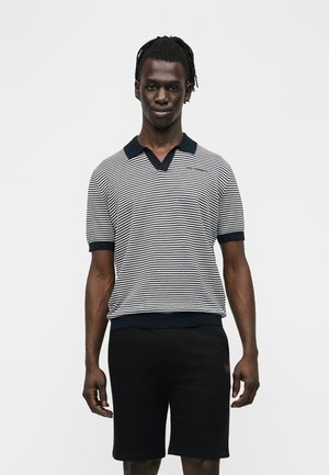 Homme portant un polo à manches courtes rayé noir et blanc et un short noir, debout devant un fond clair uni.