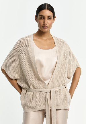 Frau trägt beige offene Strickjacke mit Gürtel über einem hellen Satin-Top und Hose, steht mit Händen in den Hüften vor schlichten Hintergrund.