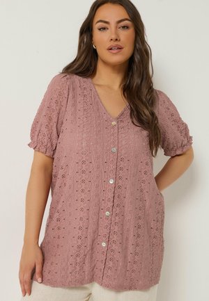 Femme portant un chemisier mauve à manches courtes avec un motif en dentelle ajourée et des boutons, posant avec une main sur la taille devant un fond uni.