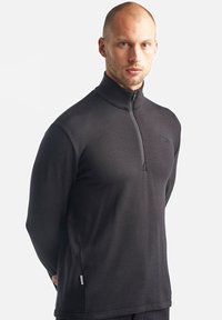 Sort fleece pullover med høj krave og halvlange lynlås. Har et diskret logo på brystet og sideslids for komfort. Glat tekstur.