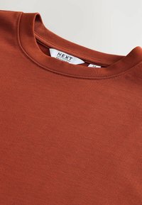 Sudadera de color óxido hecha de tejido texturizado con cuello redondo. Presenta una pequeña etiqueta de la marca en el cuello. Diseño suave y casual.