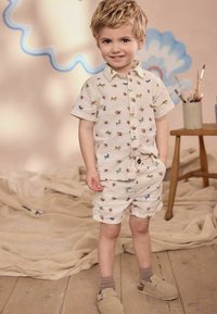 Romper a maniche corte color crema con motivi multicolori di animali e fiori, dotato di una chiusura con bottoni sul davanti e tasche laterali, abbinato a scarpe beige.