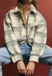 Crème en grijs geruite oversized flanellen jas met knoopsluiting en twee voorzakken, gedragen over een witte top en lichte spijkerbroek.
