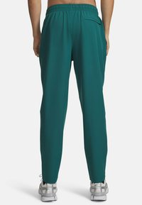 Atletske hlače v modro-zeleni barvi z elastičnim pasom in enim zadnjim žepom. Lahka tkanina z gladko teksturo. Vidni so srebrni tenis copati.