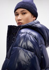 Jachetă puffer bleu marin cu o textură strălucitoare, matlasată; dispune de o glugă cu șnur și o căciulă din tricot ribat întunecată.