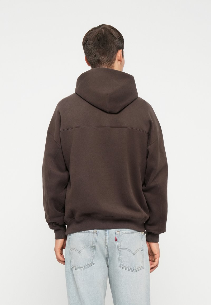 Brauner Kapuzenpullover mit lockerer Passform, weichem Material, gerippten Bündchen und breitem Bund, kombiniert mit hellblauen Jeans.