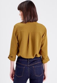 Blouse jaune moutarde avec tissu texturé, manches retroussées et détail de pli au dos. Associée à un jean foncé ajusté.