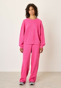 Svijetlo roza oversize dukserica s raglan rukavima, uparen s odgovarajućim širokim hlačama. Mekani materijal s suptilnom teksturom.
