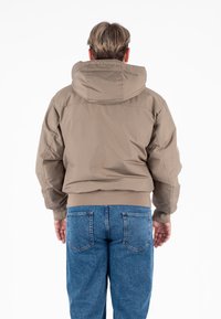 Beżowa kurtka z kapturem, zapinana na zamek, z ribbowymi mankietami, w połączeniu z niebieskimi dżinsami. Gładka tekstura, swobodny krój i lekki materiał.