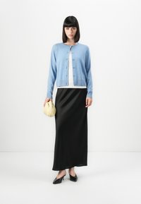Lyseblå cardigan med knappelukking, hvit topp og svart satin midi-skjørt. Holder en myk gul clutch. Svart spiss tå hæler.