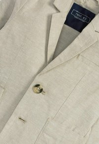 Blazer de lino beige claro con solapa con muesca, cierre de un botón y dos bolsillos frontales. Presenta una textura suave y costuras visibles.