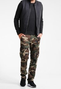 Camouflage cargo pants in green, brown, and tan with multiple pockets, paired with a black long-sleeve shirt and a gray sleeveless jacket.
