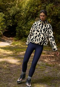 Fleece pullover vaaleassa kermanvärissä mustalla leopardikuvioinnilla, korkea kaulus, yhdistetty tummansinsisiin legginsseihin ja harmaisiin sukkiin. Ulkona vihreän ympäristön keskellä.