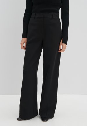 Femme portant un pantalon noir large taille haute associé à un haut noir à manches longues ajusté, debout devant un fond clair uni.