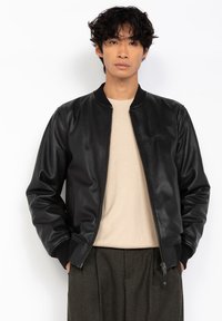 Joven con cabello rizado que lleva una chaqueta bomber de cuero negra sobre un suéter beige y pantalones oscuros, de pie contra un fondo blanco.