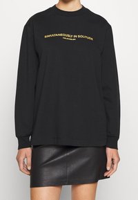 Czarna bluza z długim rękawem i okrągłym dekoltem, z żółtym napisem "JEDNOCZEŚNIE W SAMOTNOŚCI" na klatce piersiowej. Gładki materiał, luźny krój.
