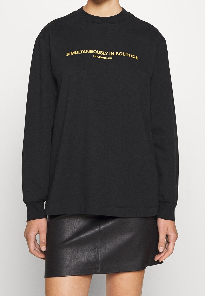 Czarna bluza z długim rękawem i okrągłym dekoltem, z żółtym napisem "JEDNOCZEŚNIE W SAMOTNOŚCI" na klatce piersiowej. Gładki materiał, luźny krój.