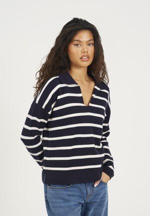 Vrouw met lang, krullend haar in een marineblauw en wit gestreept sweater en blauwe jeans, staand tegen een lichte, effen achtergrond.