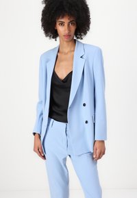 Light blue tailored suit with a double-breasted blazer and matching trousers, made of smooth fabric, worn over a black satin top.