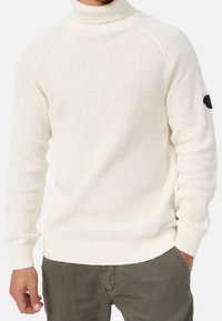 Pull à col roulé en maille blanche avec poignets et ourlet côtelés, présentant un motif texturé et une pièce de logo noire sur la manche gauche.