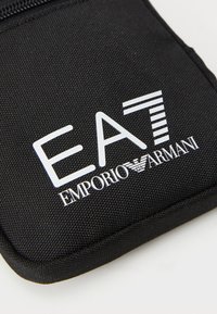 Melna auduma maiss ar baltu izdrukātu logotipu "EA7 Emporio Armani" stilizētā burtveidā un gludu rāvējslēdzēju.