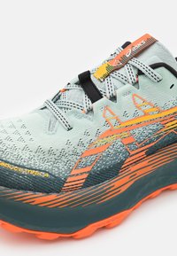Baskets de sport bleu clair avec tige en mesh, accents orange et jaune, et semelle extérieure texturée vert foncé. Présente des lacets robustes et un logo.