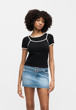 Γυναίκα με σγουρά μαλλιά που φοράει μαύρο κοντομάνικο τοπ με άσπρη επένδυση και μπλε μίνι τζιν φούστα με ζώνη, στέκεται μπροστά σε λευκό φόντο.