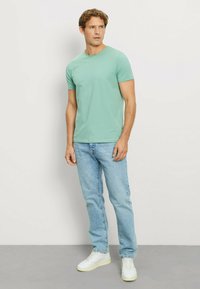 Green short-sleeve t-shirt made of soft cotton, paired with light blue jeans and white sneakers, standing against a neutral background.