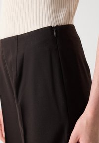 Close-up van zwarte op maat gemaakte broek met een zijrits, gedragen met een beige ribbeltrui en een hand van een persoon die naast de heup rust.