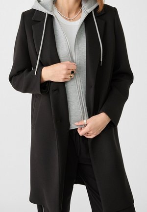 Personne portant un manteau noir par-dessus un sweat à capuche gris zippé et une chemise blanche, accessoirisée de bagues et d'un collier de perles, mains ajustant le manteau et le sweat à capuche.