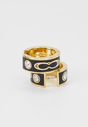 Boucles d'oreilles créoles dorées et noires avec des cristaux ronds transparents et un motif de boucle entrelacée sur la surface noire, photographiées sur fond blanc.