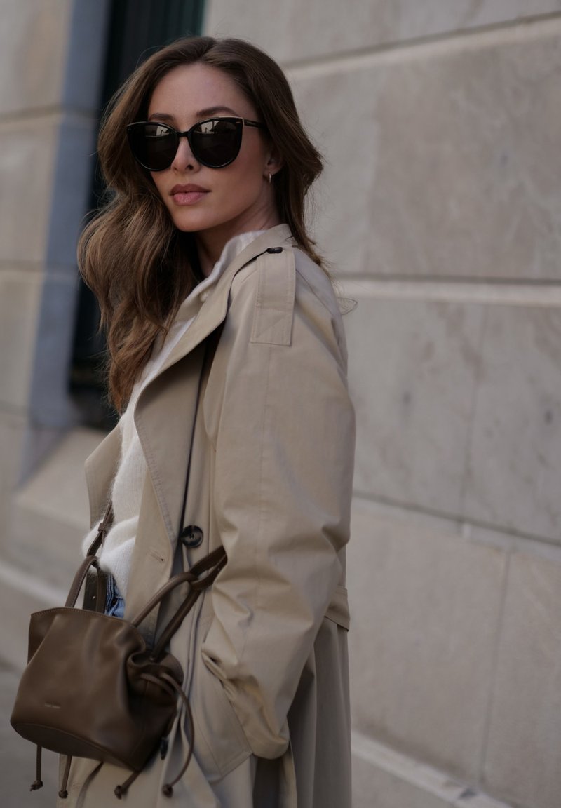 Beige trenssi takki, jossa on nappi yksityiskohtia, päällä valkoinen villapaita ja siniset farkut. Mustat aurinkolasit ja pieni ruskea laukku viimeistelevät ilmeen.