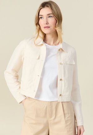 Blonde Frau, die eine hellbeige Jacke mit Knöpfen über einem weißen T-Shirt und beige Hosen trägt, steht mit einer Hand in der Tasche vor einem einfarbigen Hintergrund.