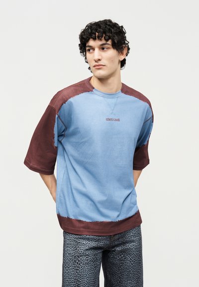 Νεαρός άνδρας με σγουρά μαλλιά φοράει φαρδιά κοντομάνικη μπλούζα μπλε και καφέ χρώματος και γκρι παντελόνι με λεοπάρ σχέδιο, κοιτάζοντας προς τα πλάγια.