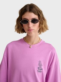 Młoda kobieta z krótkimi falowanymi włosami nosi owalne czarne okulary przeciwsłoneczne, grubą złotą łańcuchową naszyjnik z serduszkowym wisiorkiem i różową bluzę marki Hummel.