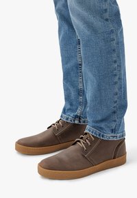 Scarpe in pelle marrone con punta rotonda, design con lacci e suola in gomma, abbinate a jeans di denim azzurro chiaro. Dettagli di cucitura sottili.