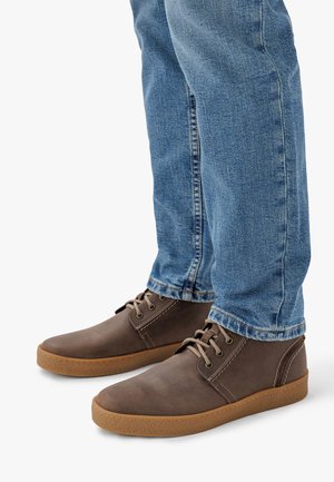 Scarpe in pelle marrone con punta rotonda, design con lacci e suola in gomma, abbinate a jeans di denim azzurro chiaro. Dettagli di cucitura sottili.