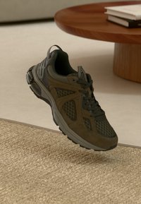 Prune ja must sportlik toss, millel on võrgust ja seemisnaha detailid, hõljuv tekstuuriga beežil vaibal, puidust kohvilauda lähedal, mille peal on kuhjatud raamatud.