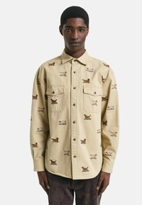 Camicia button-up beige con motivi di cani ricamati in marrone e bianco. Due tasche sul petto e un fit rilassato, realizzata in tessuto resistente.
