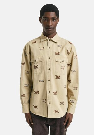 Beige overhemd met knopen, versierd met geborduurde hondenmotieven in bruin en wit. Twee borstzakken en een ontspannen pasvorm, gemaakt van stevige stof.