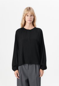 Blond kvinne iført en løs svart langermet bluse med knapper ved kragen og grå vide bukser, stående mot en hvit bakgrunn.
